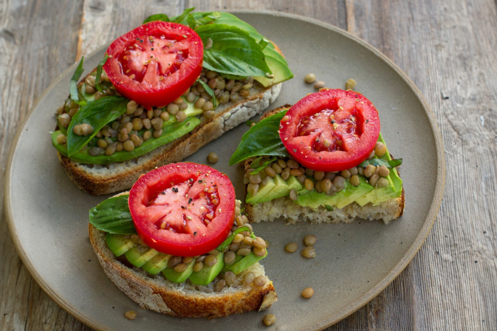 Das Bild zeigt ein Beispiel für gesunde Lunch-Pakete: zwei Brotscheiben belegt mit Avocado, Linsen, Salat und Tomate.